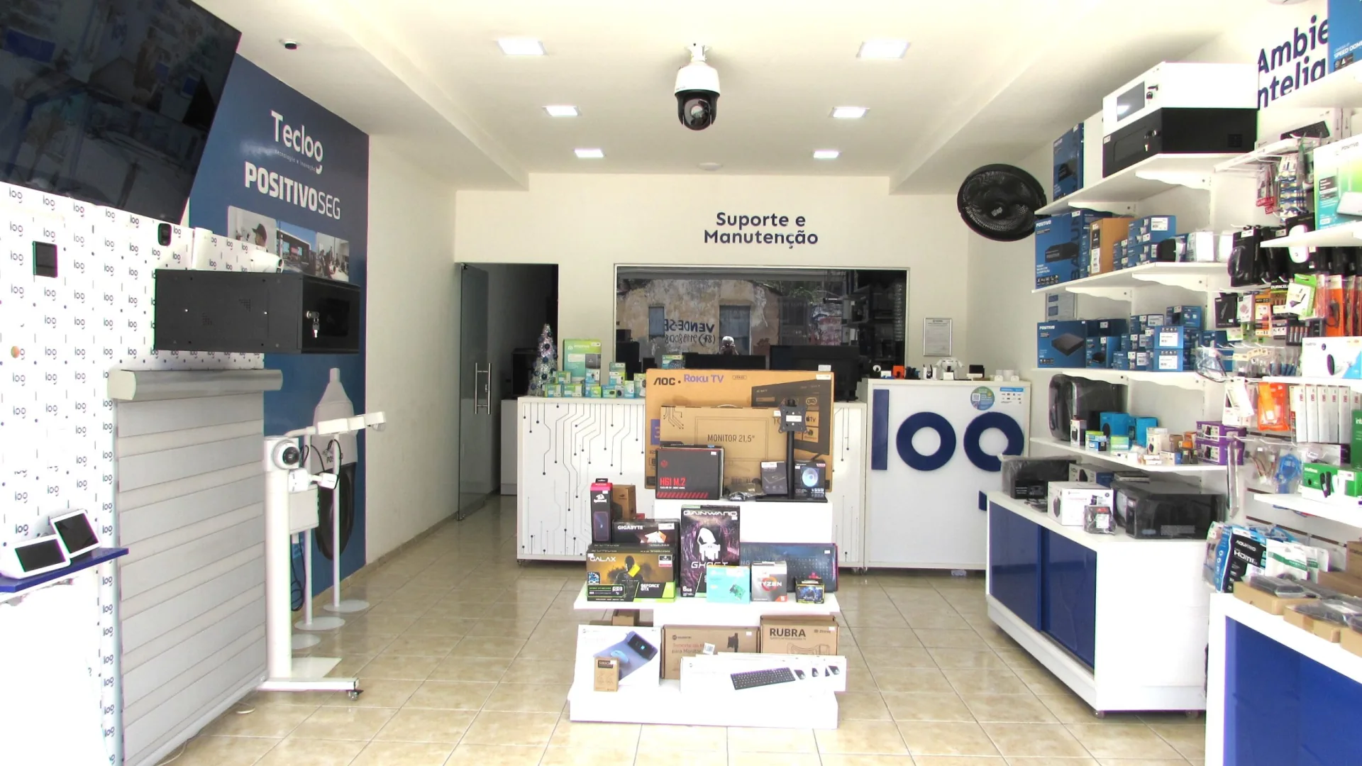 Loja Teclog vista da entrada, com prateleiras de eletrônicos, painel Positivo Seg e balcão de Suporte e Manutenção