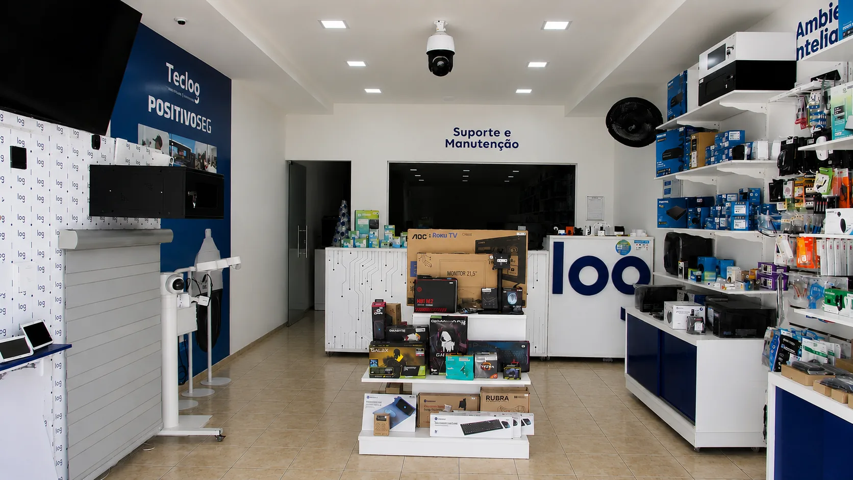 Interior da loja Teclog, com expositores de eletrônicos e câmeras de segurança, painel Positivo Seg e balcão de Suporte e Manutenção ao fundo